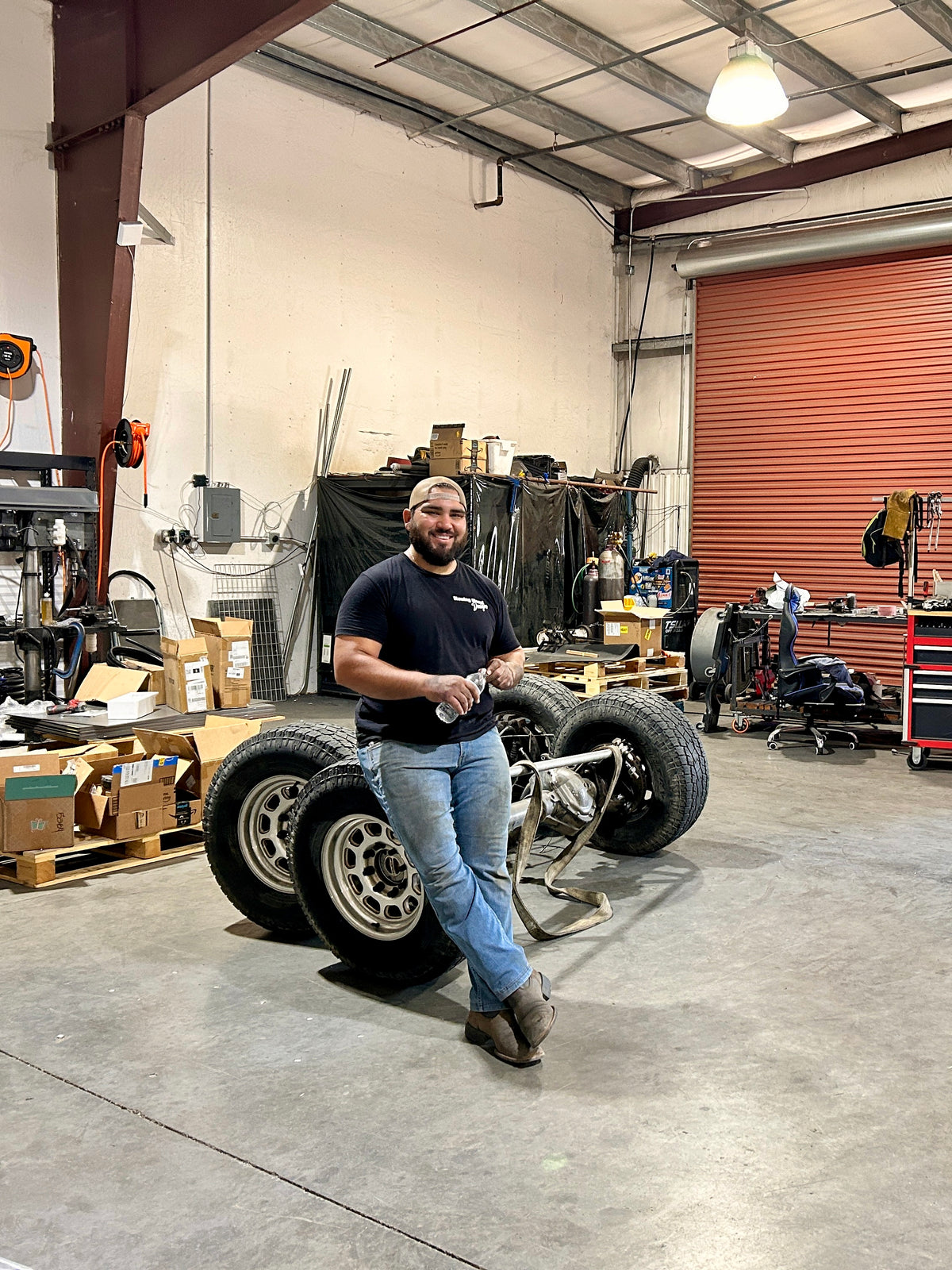 Joel Dominguez wearing a black BDD t-shirt and blue jeans stands in BDD workshop next to front and rear 2005+ axles. 