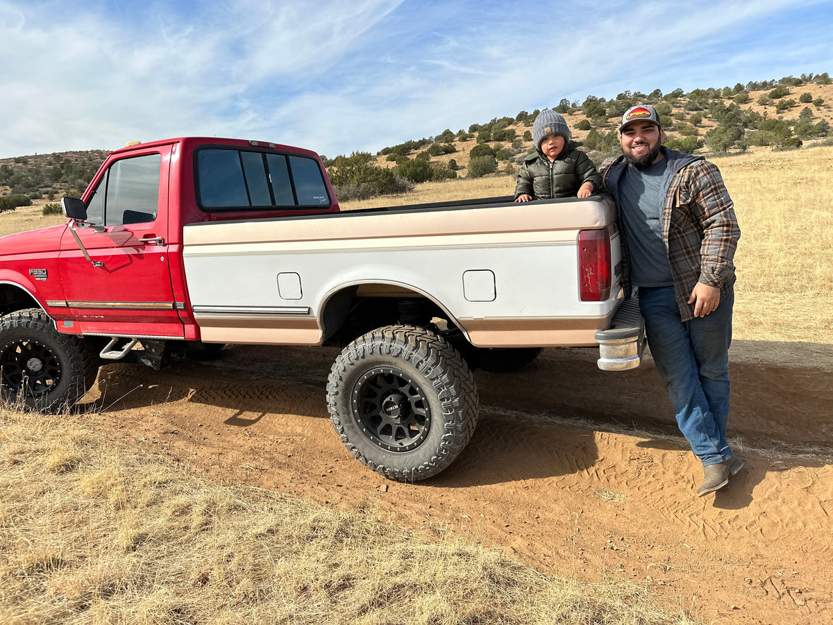 A red and beige vintage OBS pickup truck parked on a dirt road in an outdoor field, with Joel and his son beside the truck bed under a blue sky.