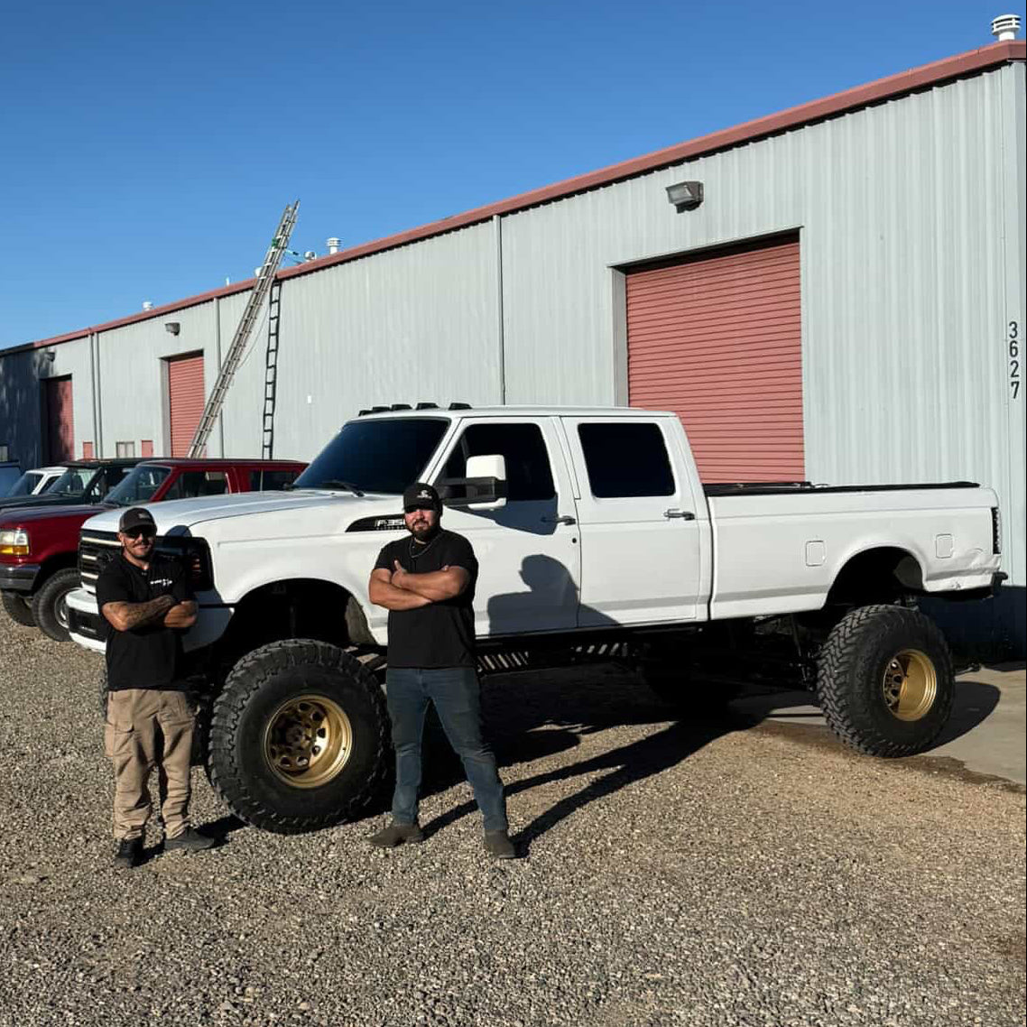 A white lifted OBS truck with large off-road tires and gold-rimmed wheels parked outdoors on gravel in front of a light gray metal BDD warehouse building under a clear blue sky. Joel Dominguez & Bryce Foster in front of finished BDD 2005+ axle swap 4-link cradle rear air ride OBS truck