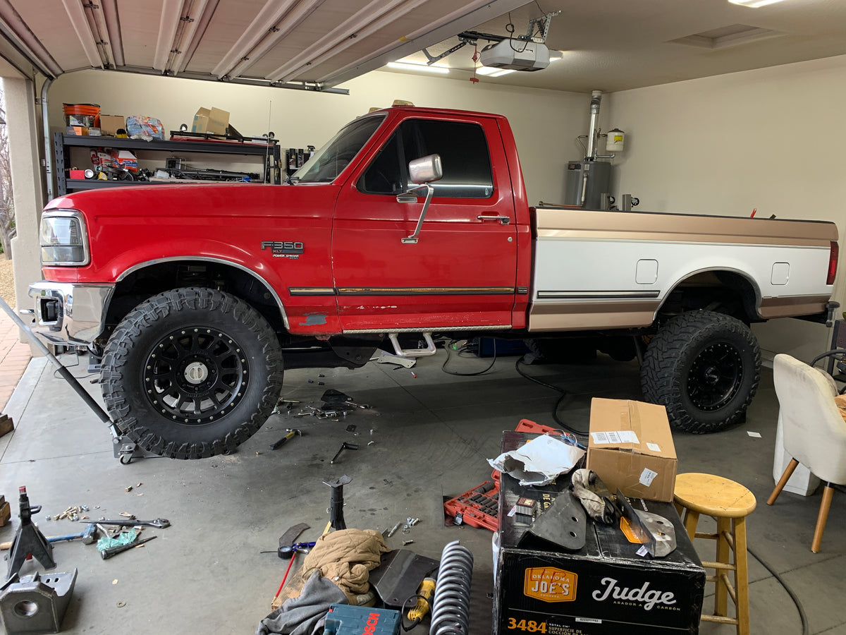 The original OBS truck that started BDD painted in red and white. It's a lifted Ford pickup truck with large off-road tires parked inside Joel's garage. The truck is raised on a jack, with tools and equipment visible on the garage floor and shelves in the background. The mess displays the work Joel was doing to develop the original BDD 2005+ axle swap kit.