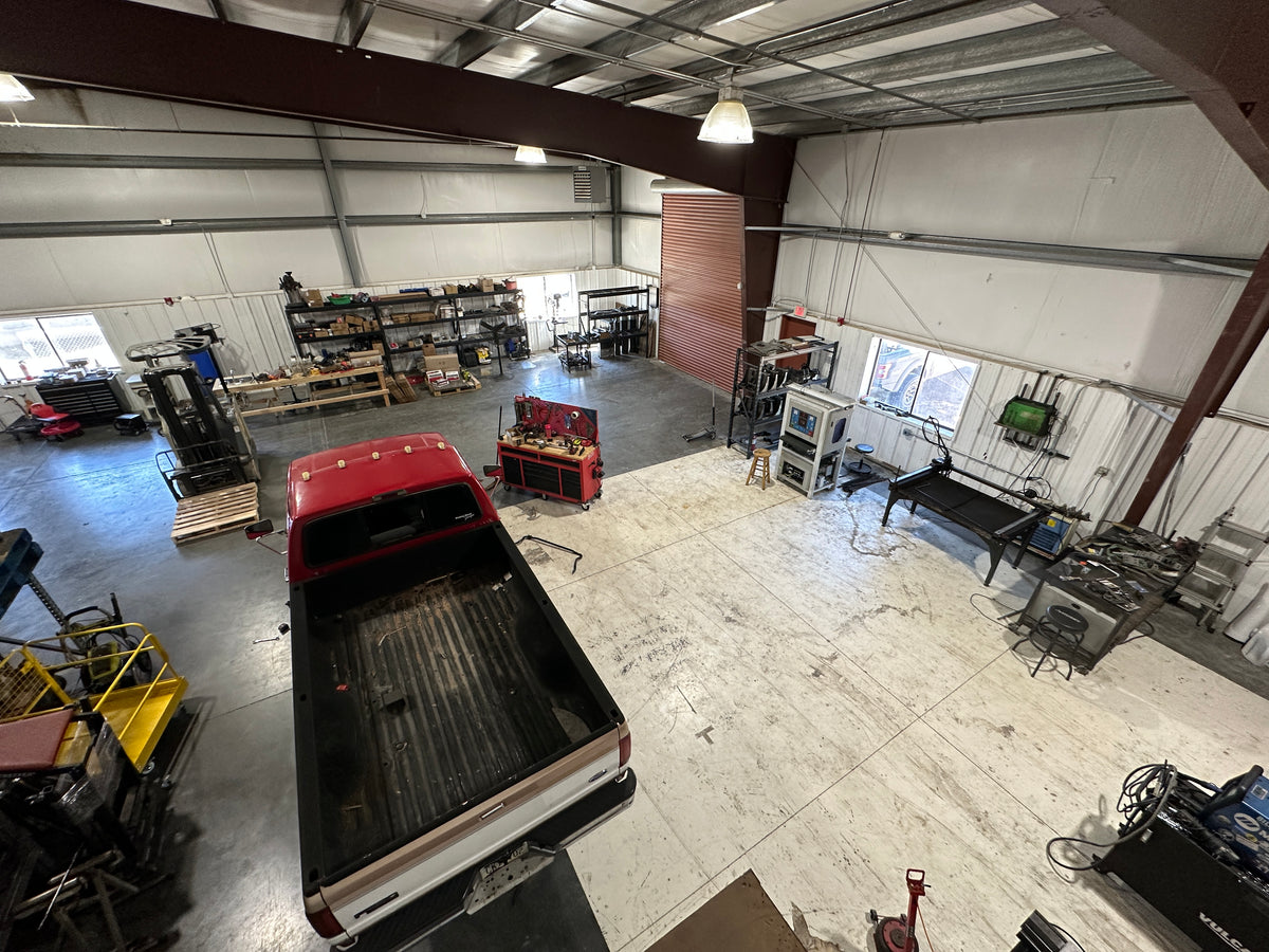 A high-angle view of a large workshop interior with a red pickup truck parked inside, surrounded by tools, equipment, shelves, and workbenches. The second BDD shop showing move in day.