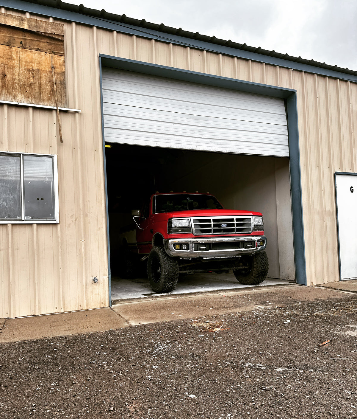 A red lifted pickup truck with large off-road tires drives out of a white roll-up garage door in a beige metal-sided warehouse or shed. The first BDD shop. 